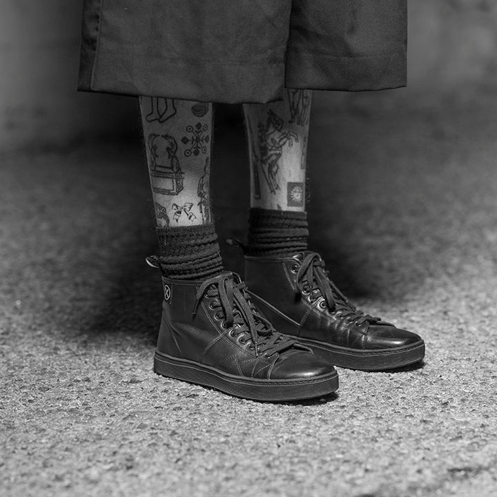 Lifestyle photo of a person wearing the CNTRL high-top black leather shoes by FACT., showing the shoes paired with socks and cropped pants on pavement.