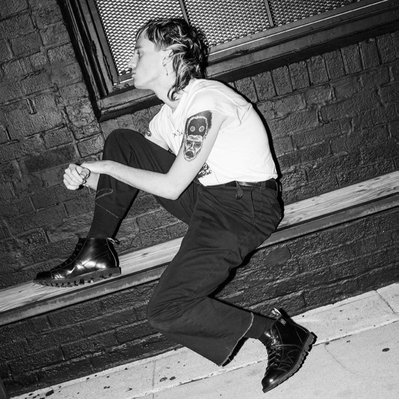 Black-and-white photo of a person with tattoos wearing MNKY boots while sitting on an urban ledge, capturing a rebellious post-punk style.