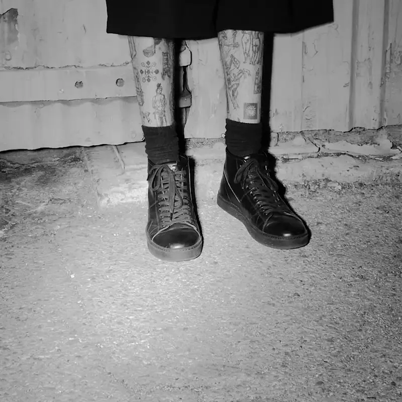 Black-and-white photo of a person standing in FACT. CNTRL high-top shoes with tattooed legs, highlighting the shoes against an industrial backdrop.