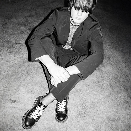 Person wearing a dark suit and black Monkey Boot )MNKY SHOE) sitting on a textured floor.
