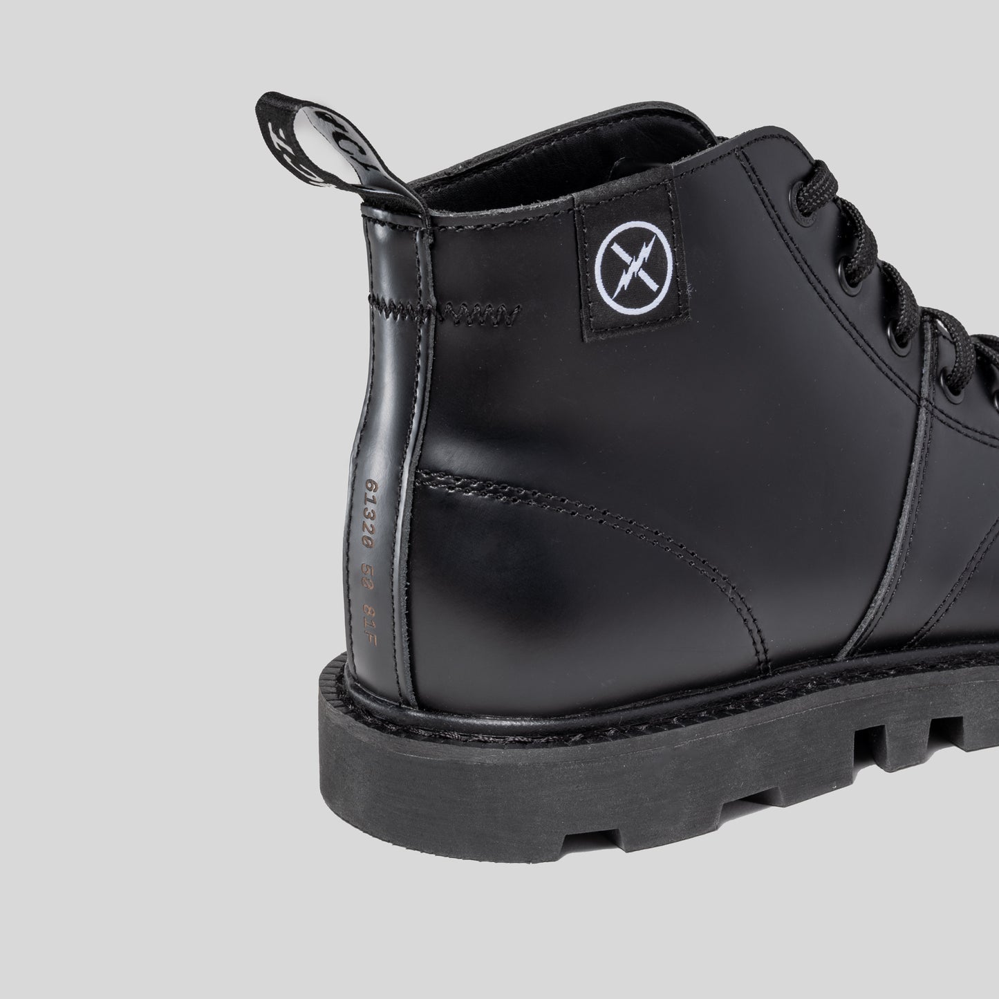 Close-up of the MNKY boot heel showing FACT. logo patch, woven heel pull tab, and welted construction on a gray background.